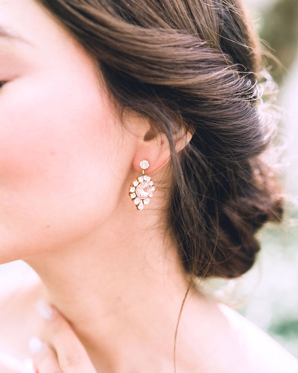 A close model view of a bride wearing the Enchanted Crystal Drop Earrings in gold with blush crystal centers. Styled with a low bridal updo with twists.