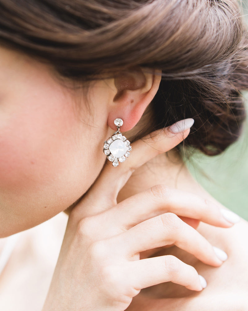 A close model view of a bride wearing the Enchanted Crystal Drop Earrings in silver with white opal centers. 