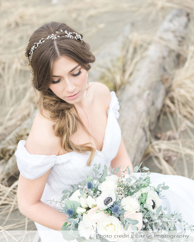 A bride sits gracefully on a log at the beach. Her hair is styled down and to the side. She wears the Everthine Crystal Hair Vine in the front.