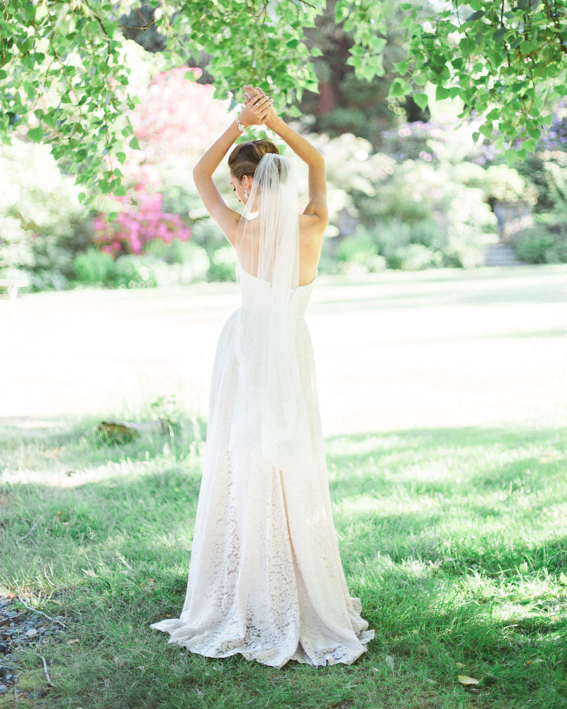A model stands with her back to the camera in a sunlit field. She wears the Flora Veil with Alencon lace border.