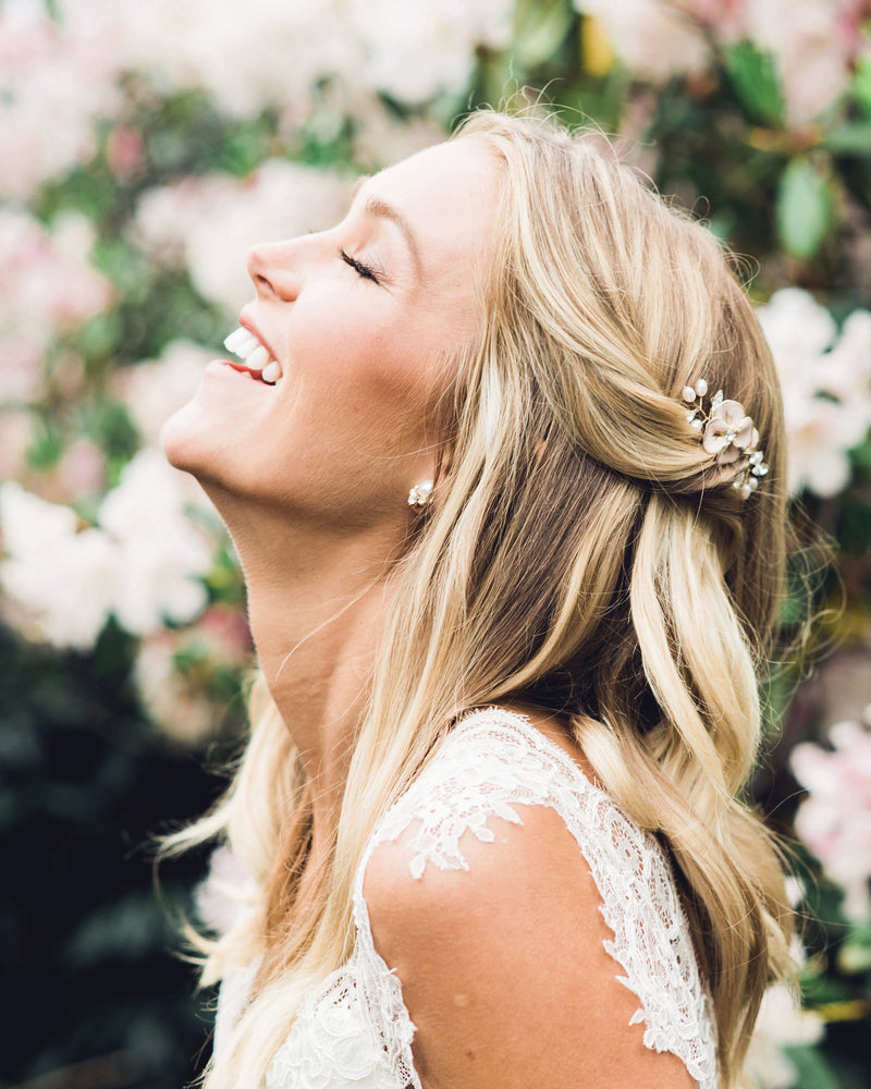 A model laughs happily as she models a bridal comb. Her hair is styled in soft waves with both sides pinned back. She wears the Gilded Blossoms Petite Comb styled into the side. The comb has blush hand-painted flowers, pearls, and crystals.
