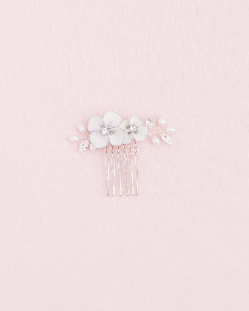 Flatlay on a pink background of the Gilded Blossoms Petite Comb in silver. The comb has white flowers hand-painted with a silver edge, pearls, and crystals.