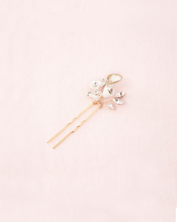 Flatlay on pink background of the Gilded Lily Hair Pin in gold. The hair pin has a white lily flower with gold hand-painted edges, smaller white flowers, and scattered crystals.