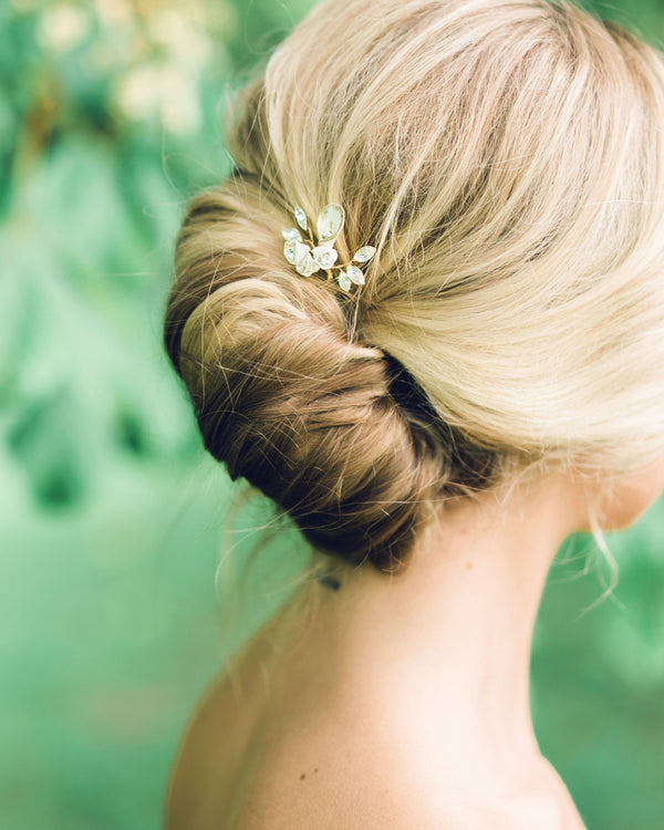 A blonde model has her hair styled into a modern bridal French twist. The Gilded Lily Hair Pin is styled into the updo. The hair pin has white flowers and crystals.