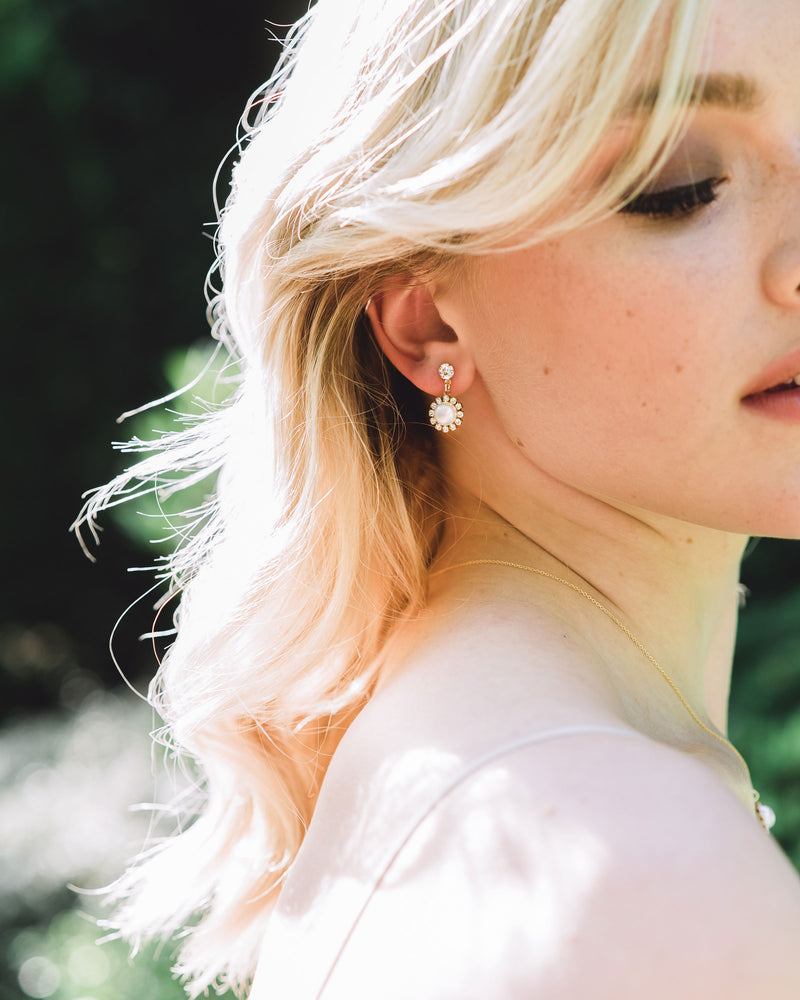 A blonde model with her hair styled in with soft bridal waves is wearing the Halo Pearl Drop Earrings in gold with natural freshwater pearls.
