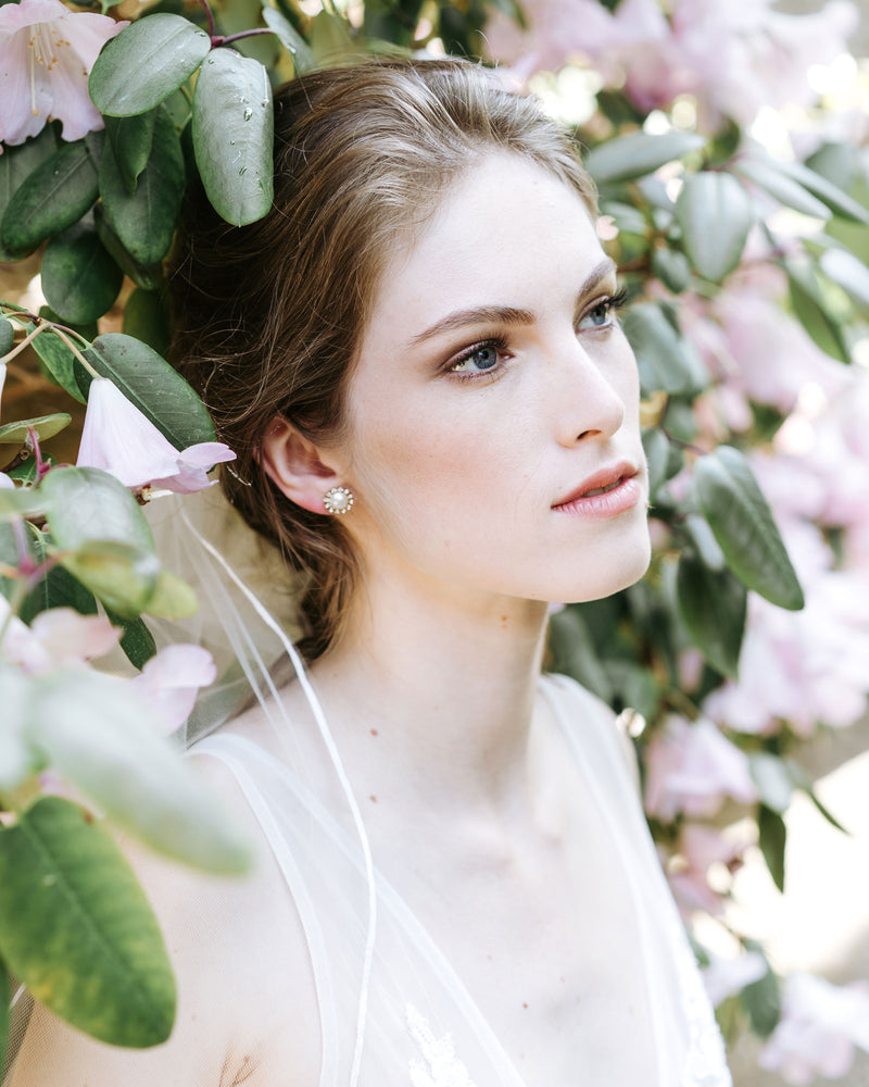 A model is surrounded by flowering trees. She wears the Halo Pearl Stud Earrings with the Leila Ribbon Veil. 