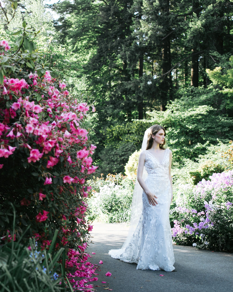 A bride stands in a flower garden. She wears a long veil with no edge.