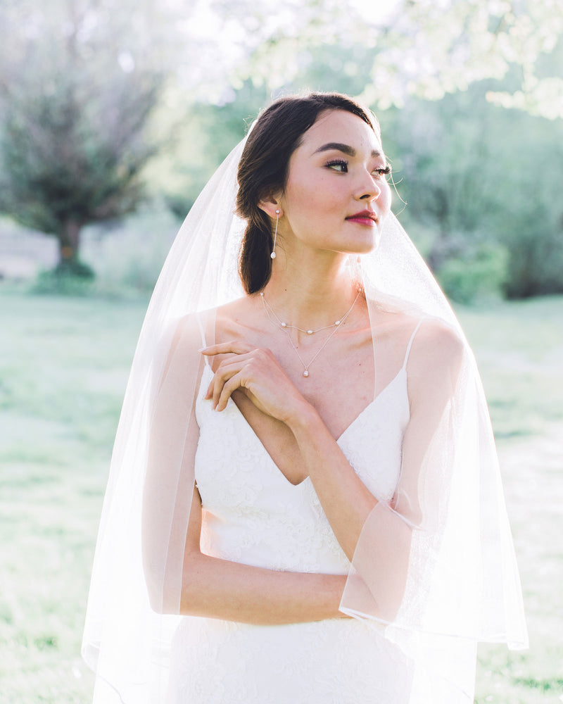 A bride wears a modern pearl jewelry set with Long Pearl Teardrop Earrings and a Layered Pearl Drop Necklace, paired with a two-layer veil with a ribbon trim.