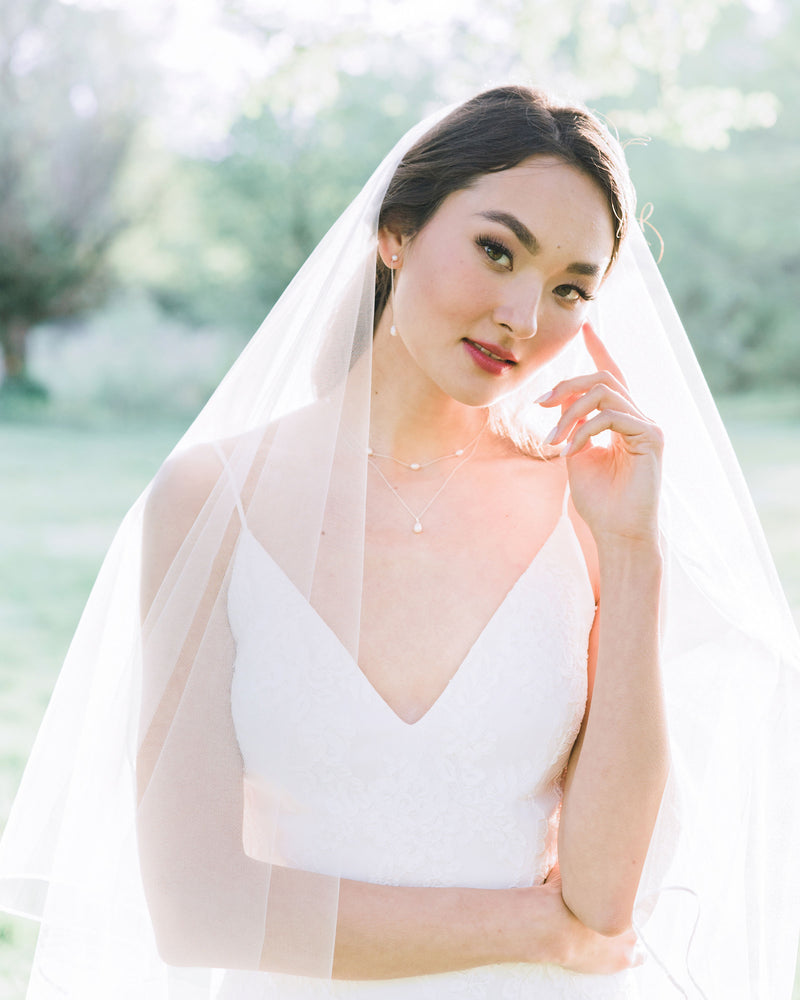 A bride wears a modern pearl jewelry set with Long Pearl Teardrop Earrings and a Layered Pearl Drop Necklace, paired with a two-layer veil with a ribbon trim.