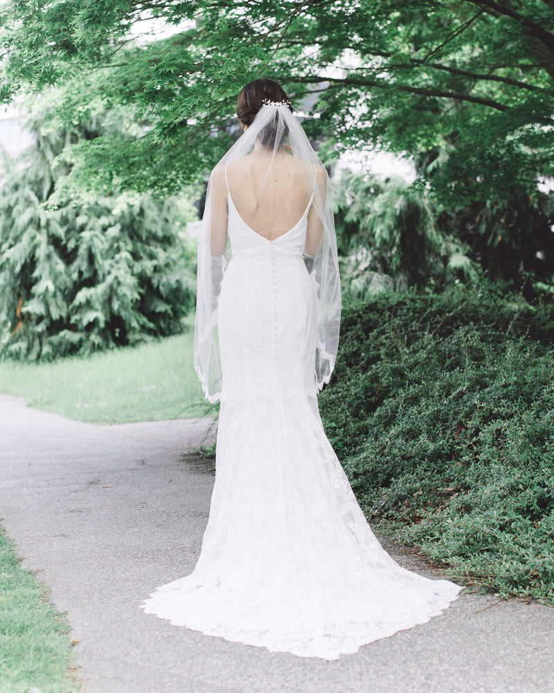 A bride models the Wisteria Lace Veil from the back; a fingertip veil with lace starting at the elbow.
